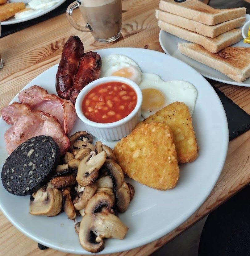 A full breakfast with bacon, sausages, black pudding, mushrooms, hash browns, fried eggs and beans, with a stack of toast and a glass of coffee behind