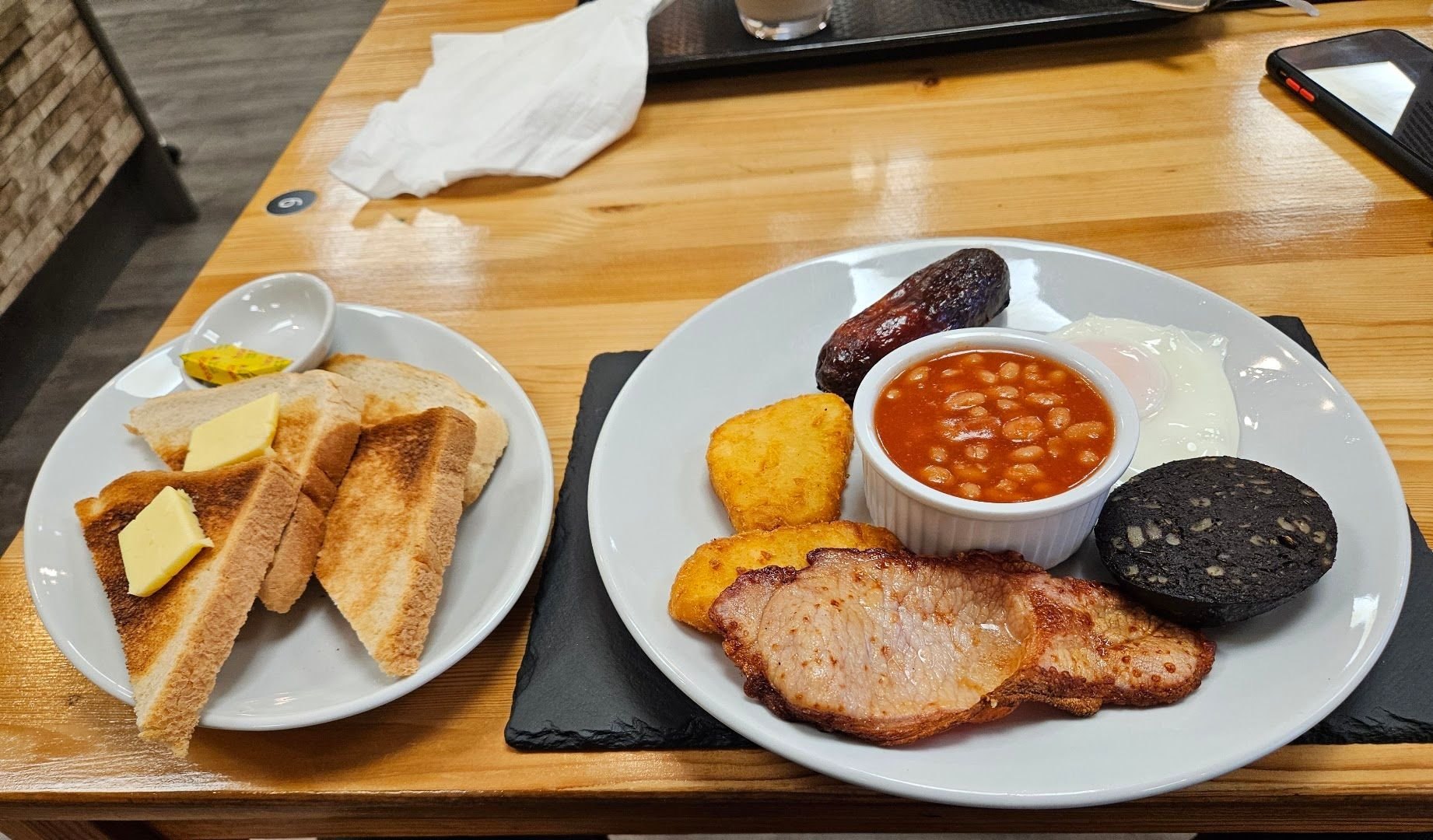 A full breakfast on a slate: bacon, sausage, black pudding, hash browns, a fried egg and a pot of beans, with buttered toast on the side